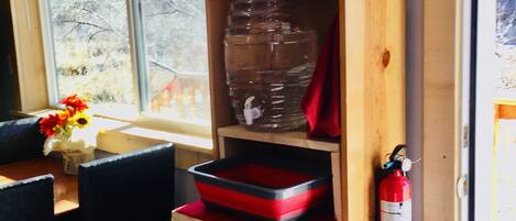 Eine Küche mit einem Holzboden, der einen Wasserspender, einen roten Mülleimer und einen roten Behälter beherbergt. Ein Fenster mit einem Karomuster-Vorhang ist sichtbar, und ein Feuerlöscher ist an der Wand montiert.