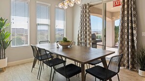 Een eethoek met een lange tafel, zwarte stoelen, een potplant en een hanglamp.