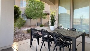 Patio esterno con tavolo e sedie, circondato da un giardino e un edificio.