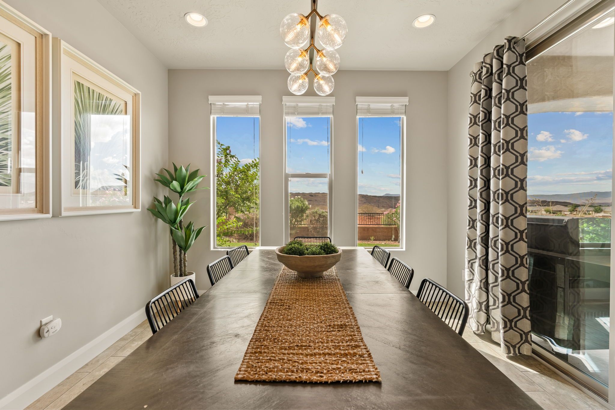 A dining area with a long table, chairs, a central plant, and large windows offering a view of the outdoors.