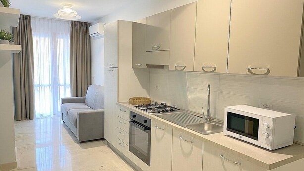 A modern kitchen with a gas stove, microwave, and stainless steel sink. There is a grey sofa and a window with curtains in the adjacent living area. - Casa Marinara C: A modern and graceful apartment situated at a short distance from the beach, with Free WI-FI. (Balestrate)