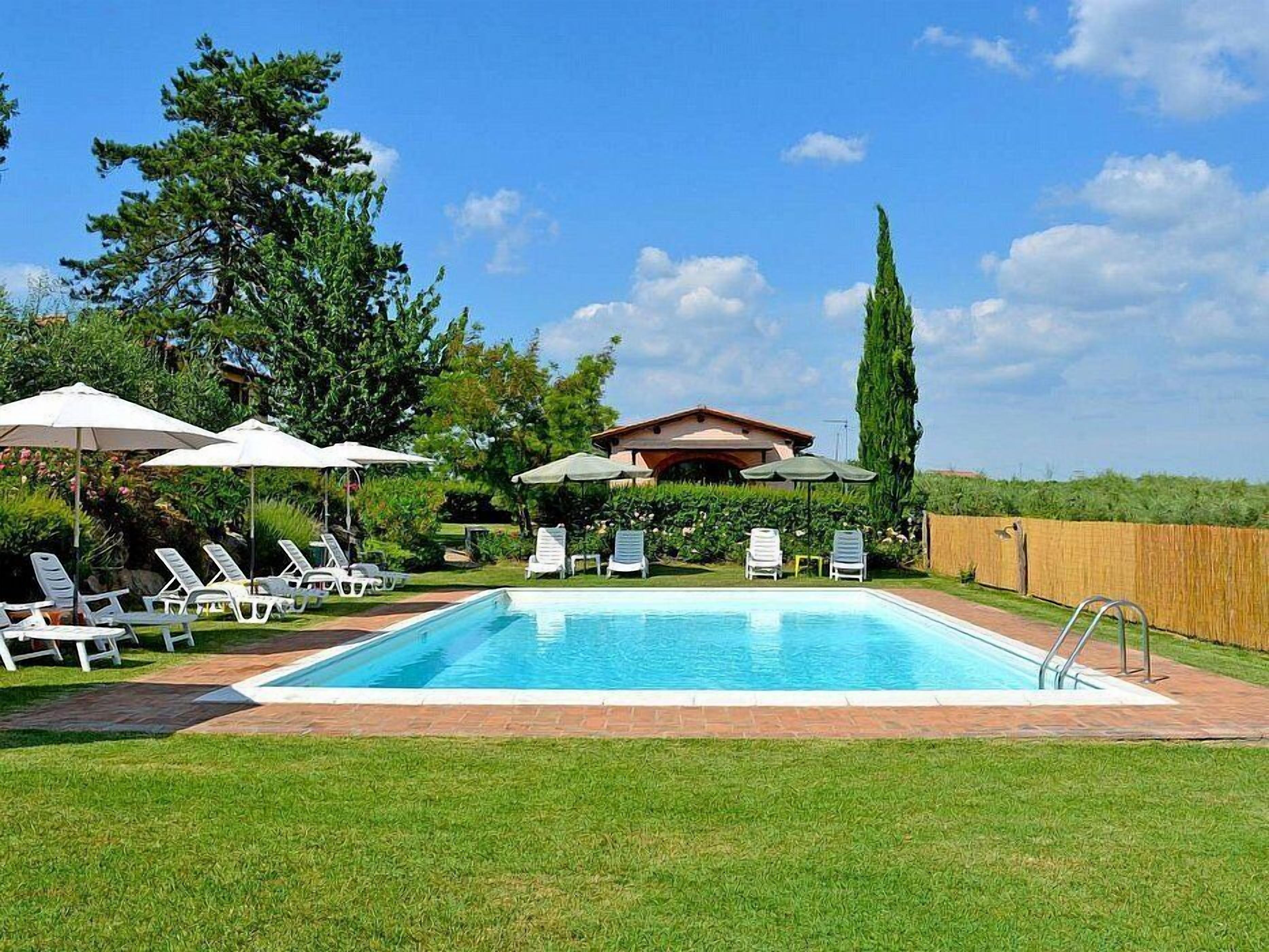 A swimming pool with lounge chairs and umbrellas, surrounded by a wooden fence and greenery.
