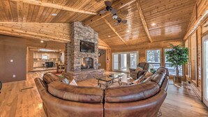 A spacious living room with a stone fireplace, leather sofa, and a large window offering a view of the outdoors. - Spacious Ruidoso Home w/ Hot Tub & Fireplaces (Ruidoso)
