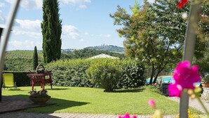 A garden with a lawn, a wooden bench, a table, and a view of a hill with buildings in the distance.