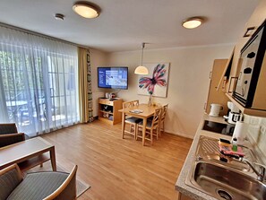 Une chambre d’hôtel moderne avec une table à manger en bois, des chaises, un téléviseur, un tableau, un coin cuisine avec un évier et une vue sur l’extérieur grâce à de grandes fenêtres.