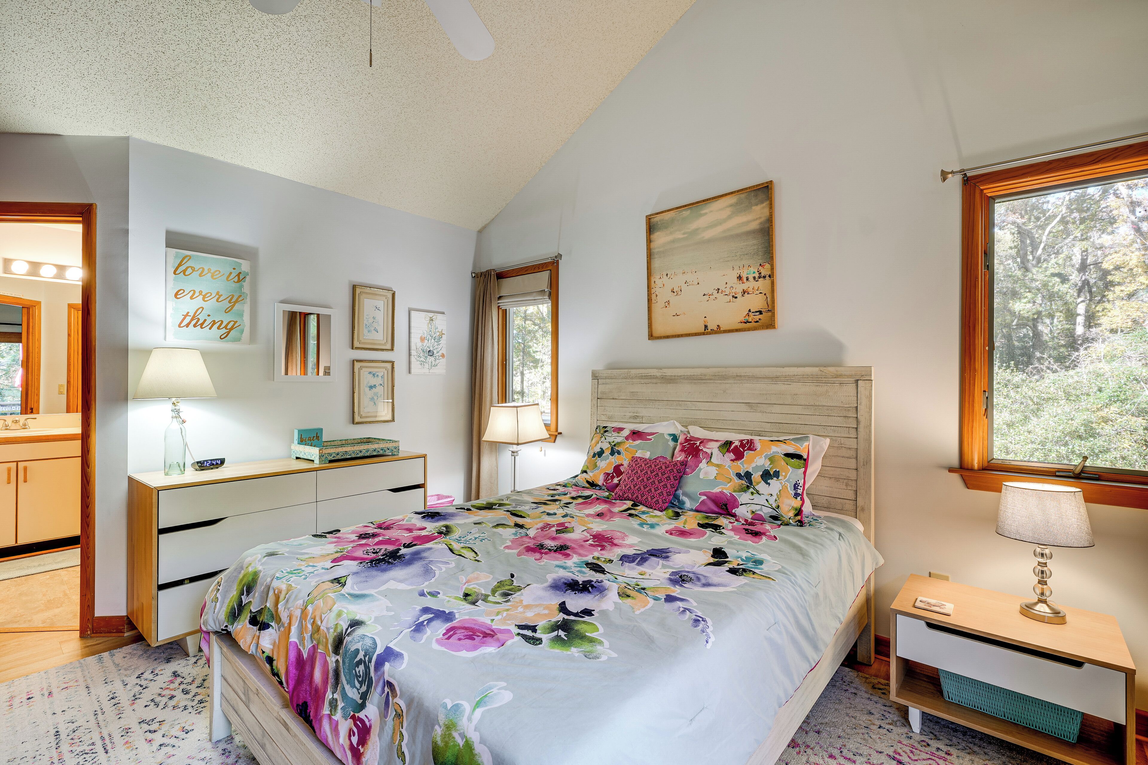 A bedroom with a floral bedspread, a wooden headboard, a nightstand with a lamp, and a window with a view of trees.