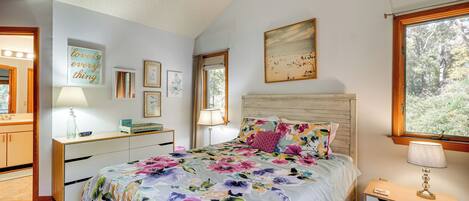 A bedroom with a floral bedspread, a wooden headboard, a nightstand with a lamp, and a window with a view of trees.