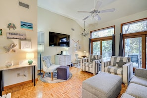 A spacious living room with a large TV, comfortable seating, a ceiling fan, and a view through large windows. - 1/2 Mi to Beach Access: Southern Shores Gem + Deck (Southern Shores)