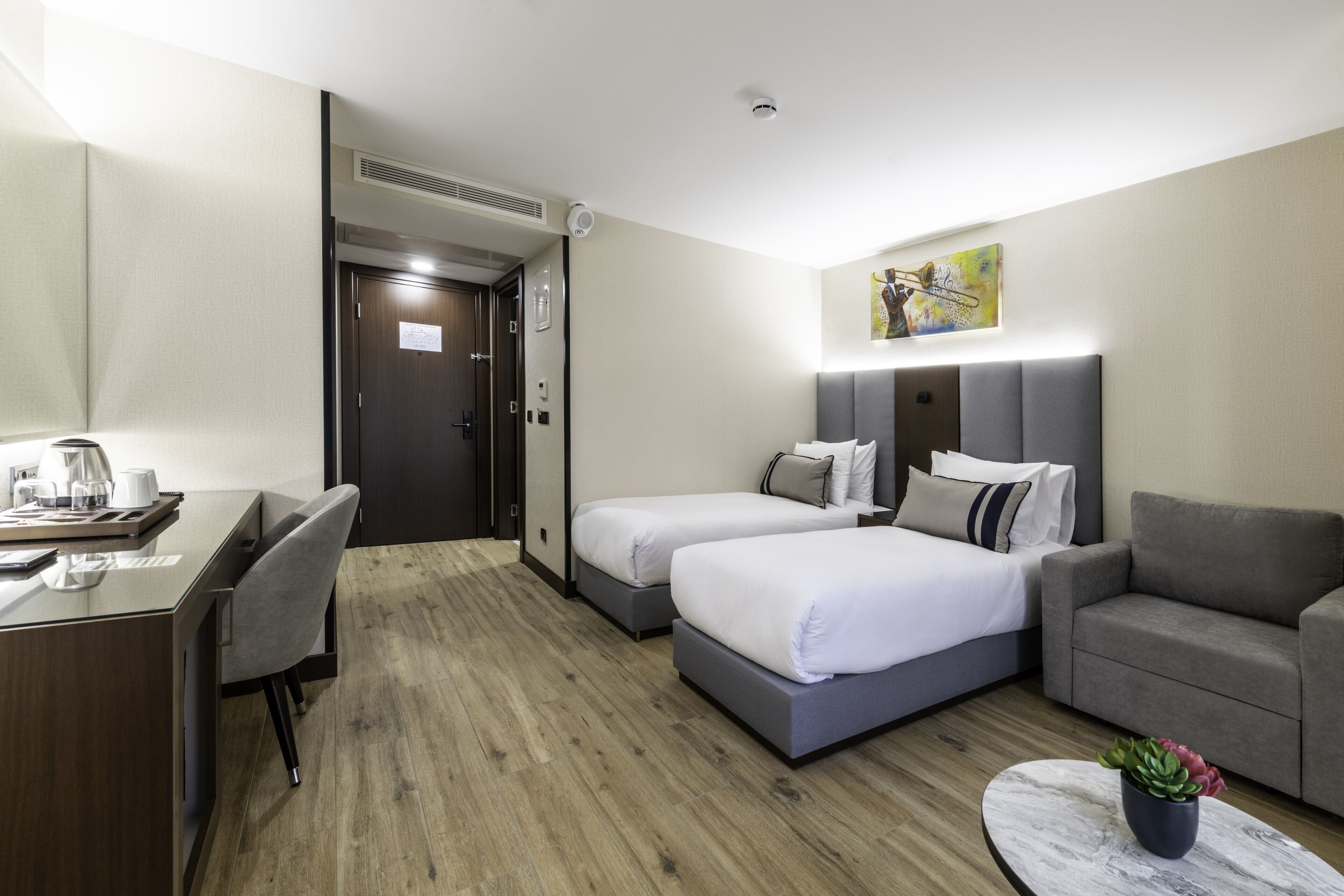 A hotel room with two beds, a desk, a chair, a sofa, and a small table with a plant.