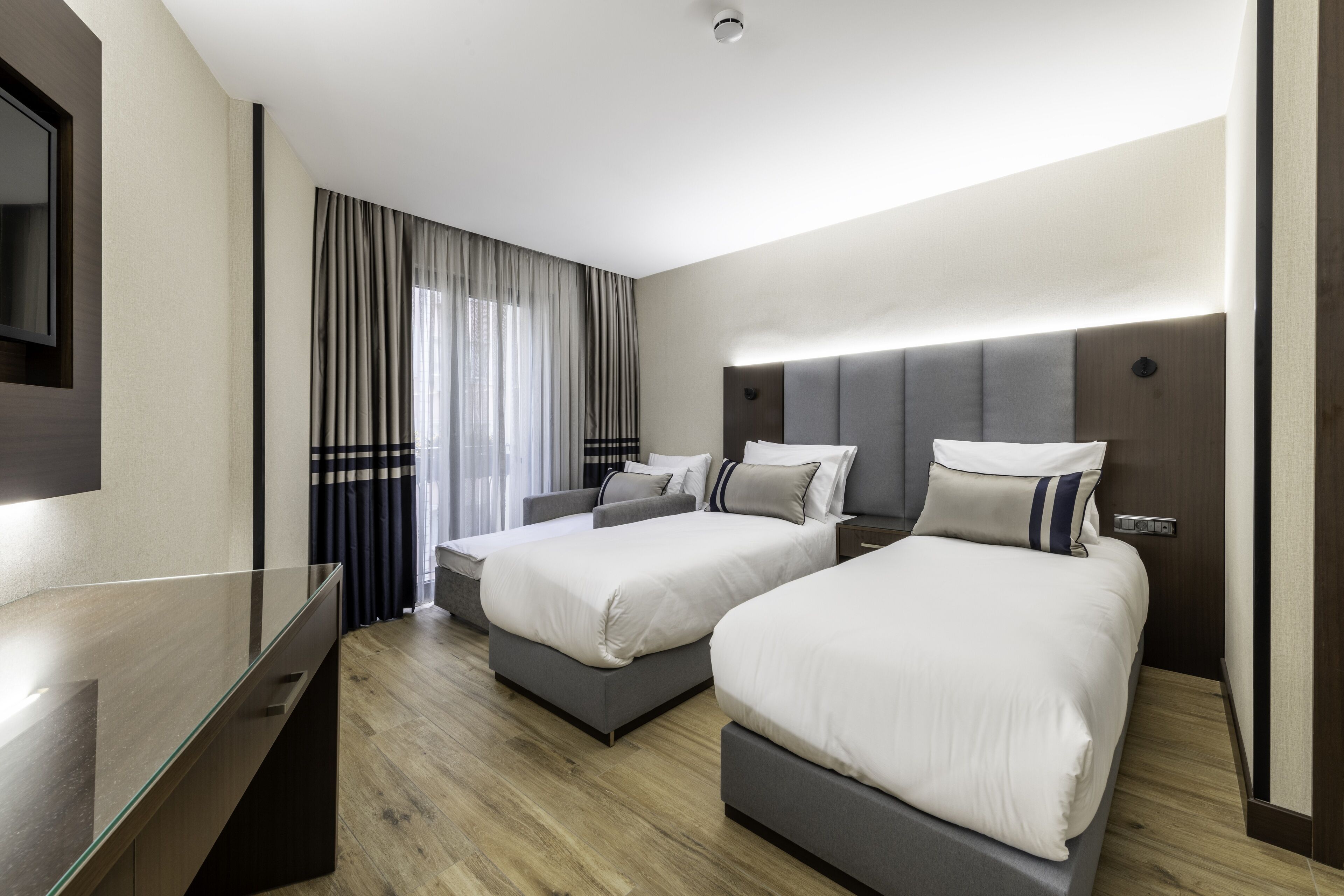 A modern hotel room with two single beds, a flat-screen TV, and a large window with curtains.