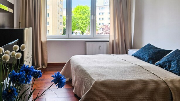 A bedroom with a bed, a window, curtains, a radiator, and a vase with flowers. - Matejki - Ma05 (Swinoujscie)