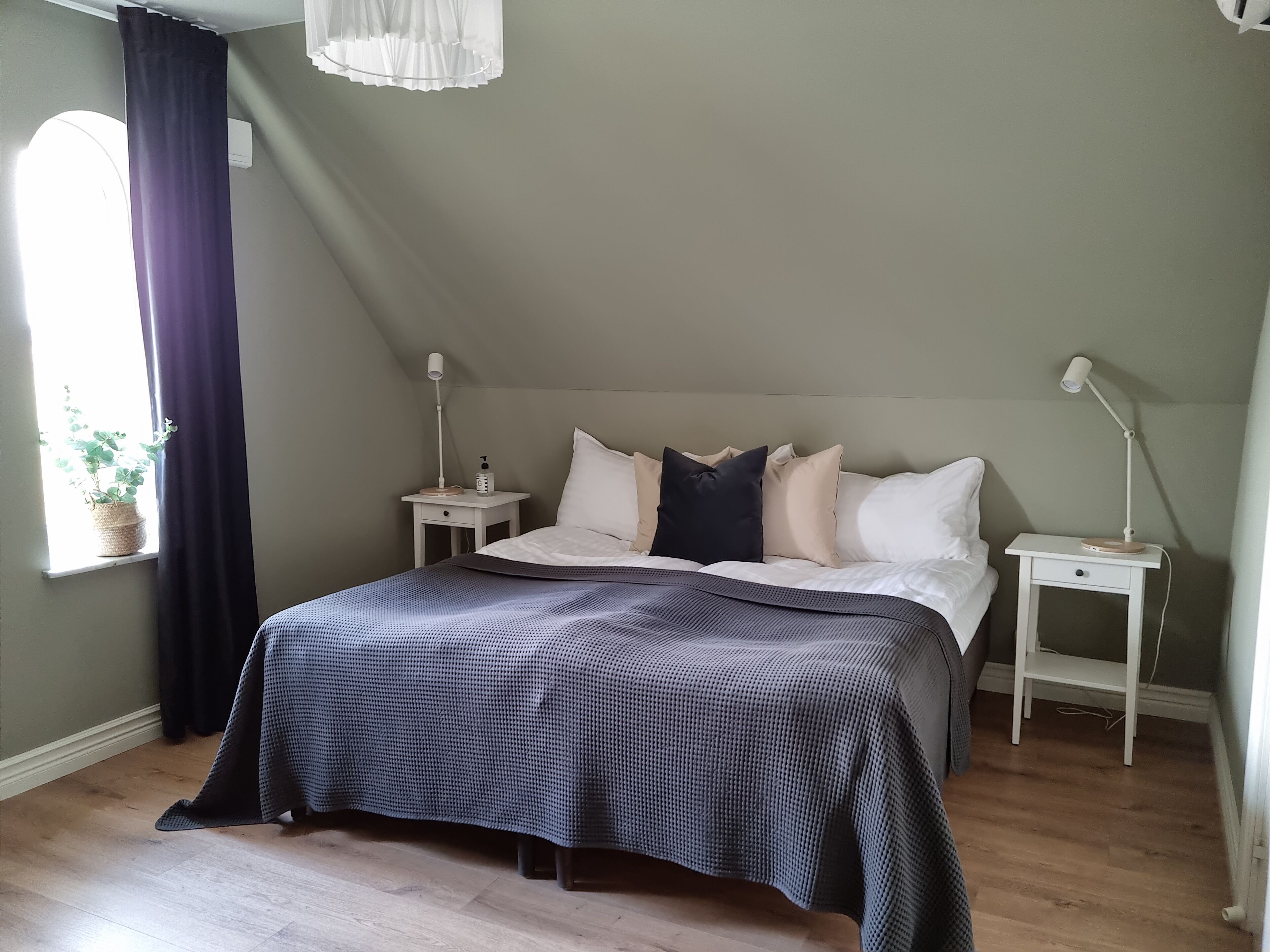Een slaapkamer met een bed, nachtkastjes, een lamp, een bloempot en een raam met gordijnen.