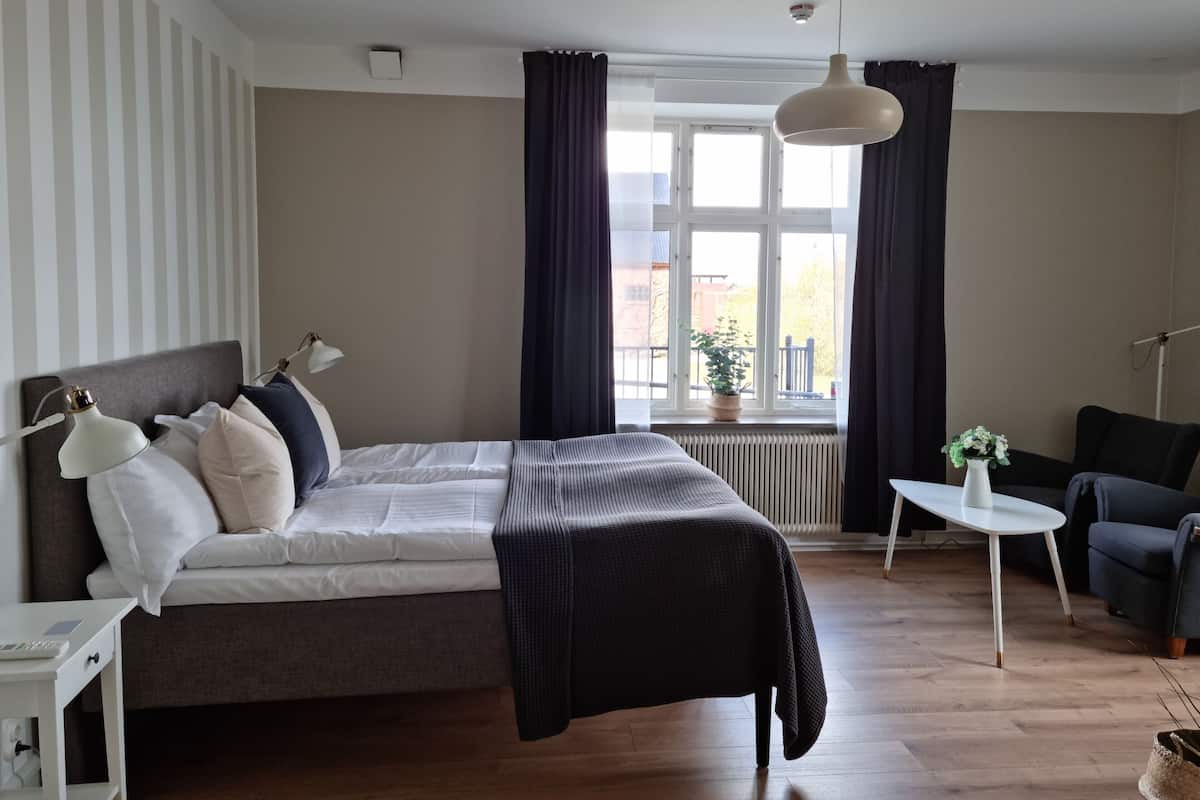 Quarto com cama, mesa de cabeceira, luminária, janela com cortinas, radiador e uma cadeira.
