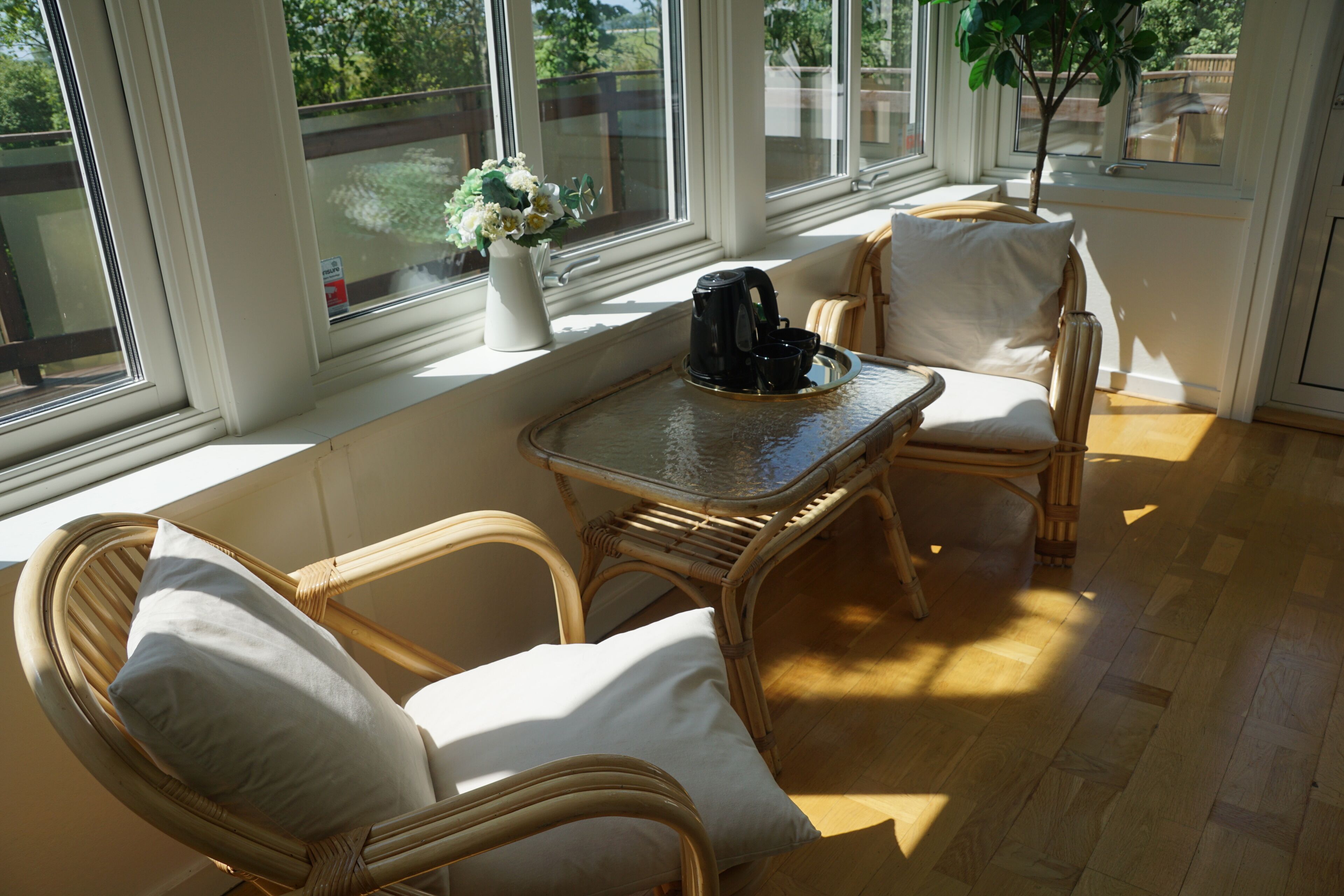 A sunlit room with wicker furniture, a coffee maker on a tray, and a window with a view of greenery.