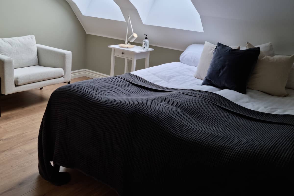 Um quarto com claraboia, mesa de cabeceira com luminária, cadeira e cama com almofadas.