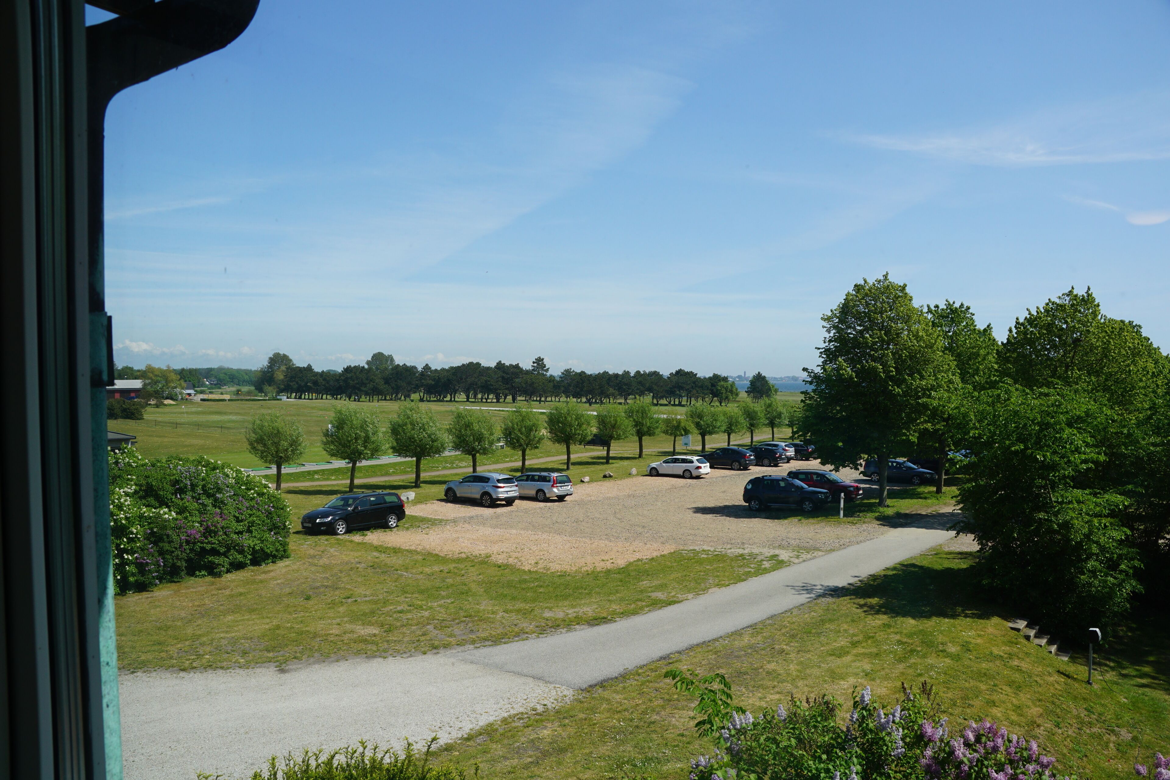 A parking area with multiple cars, a gravel path, and green trees.