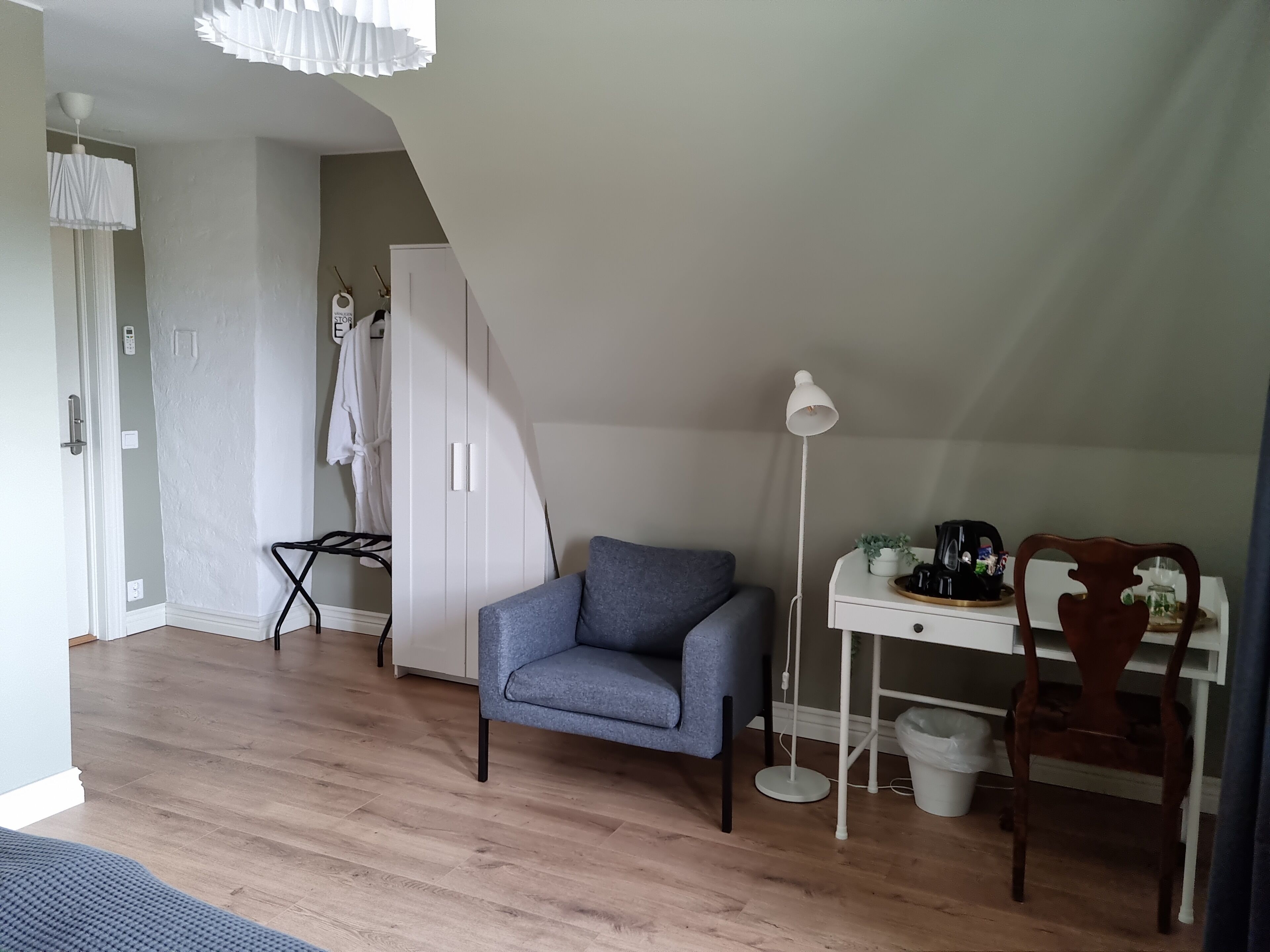 Una habitación con un sofá gris, un escritorio blanco con una lámpara, una silla de madera, un armario blanco y una planta pequeña sobre el escritorio.