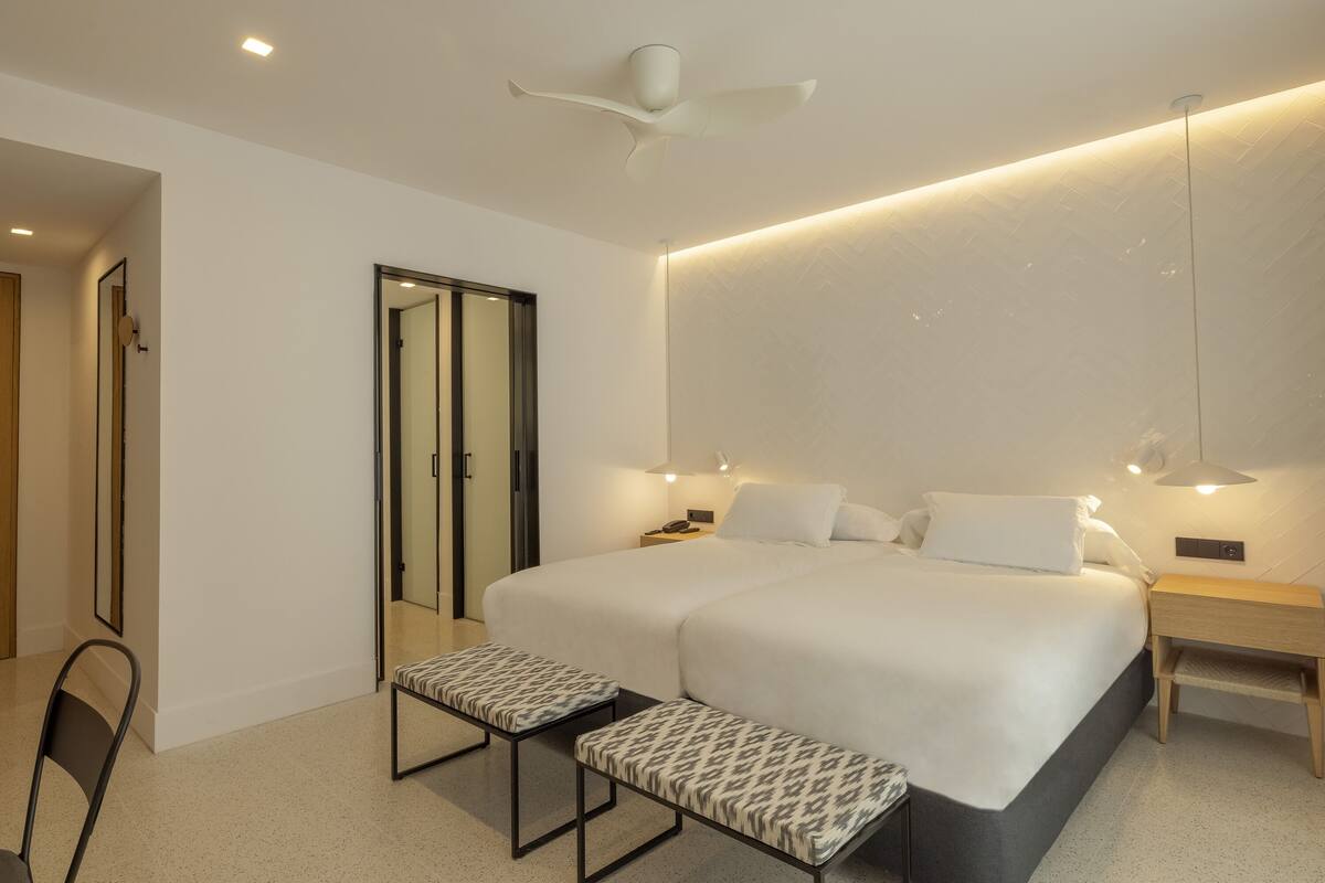 Een moderne hotelkamer met een groot bed, nachtkastjes, een bankje, een plafondventilator en een spiegel.