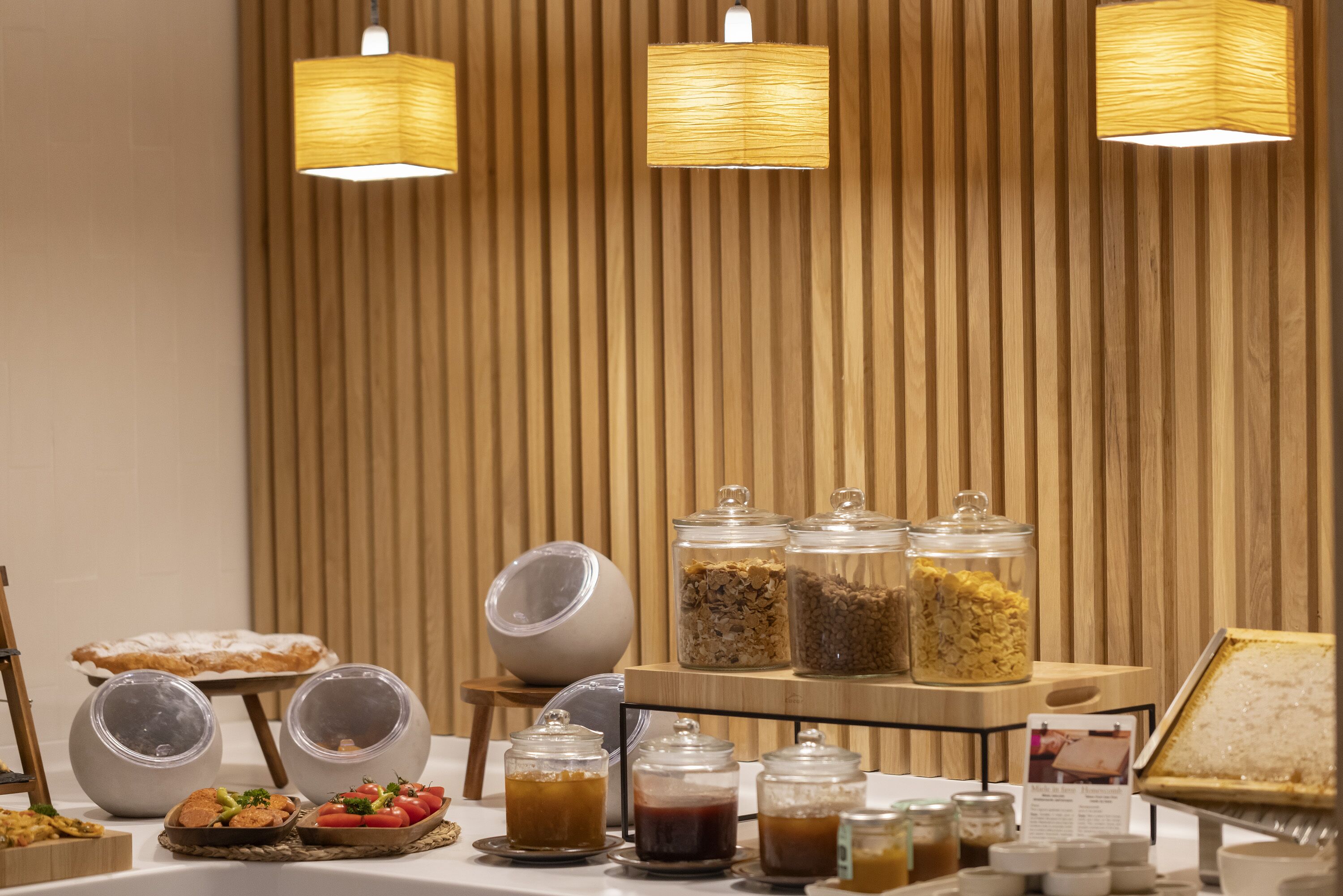 Un buffet de petit-déjeuner proposant une variété d’aliments et de boissons, notamment des céréales, des fruits et des produits laitiers, présentés sur une table avec un fond en bois et un éclairage suspendu.