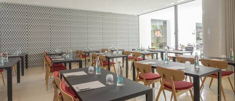 Un espace repas moderne avec des tables et des chaises, de grandes fenêtres et un mur décoré d’un motif.