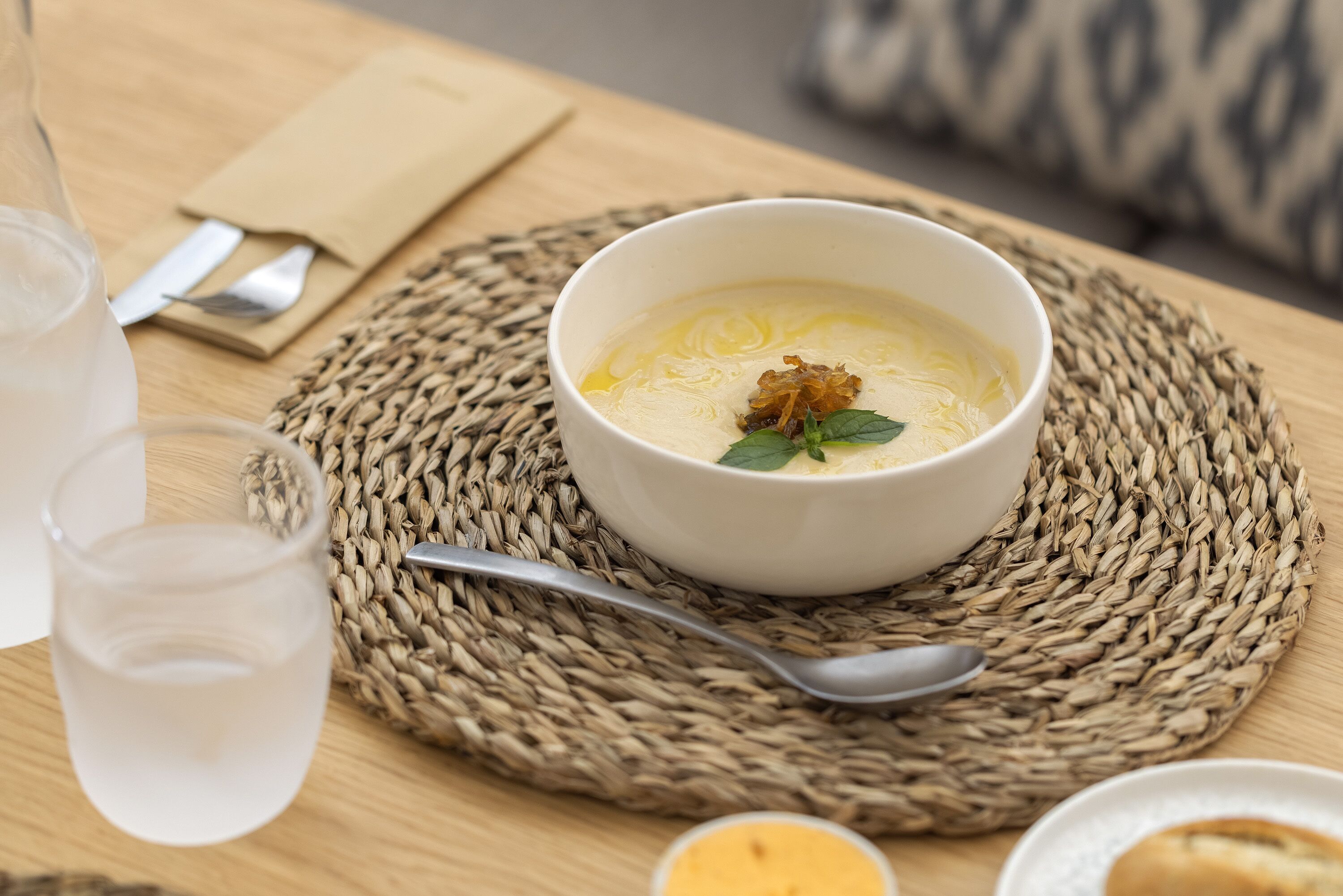 En skål suppe på en vævet bordskåner, en ske, en gaffel, en serviet og et glas vand på et træbord.