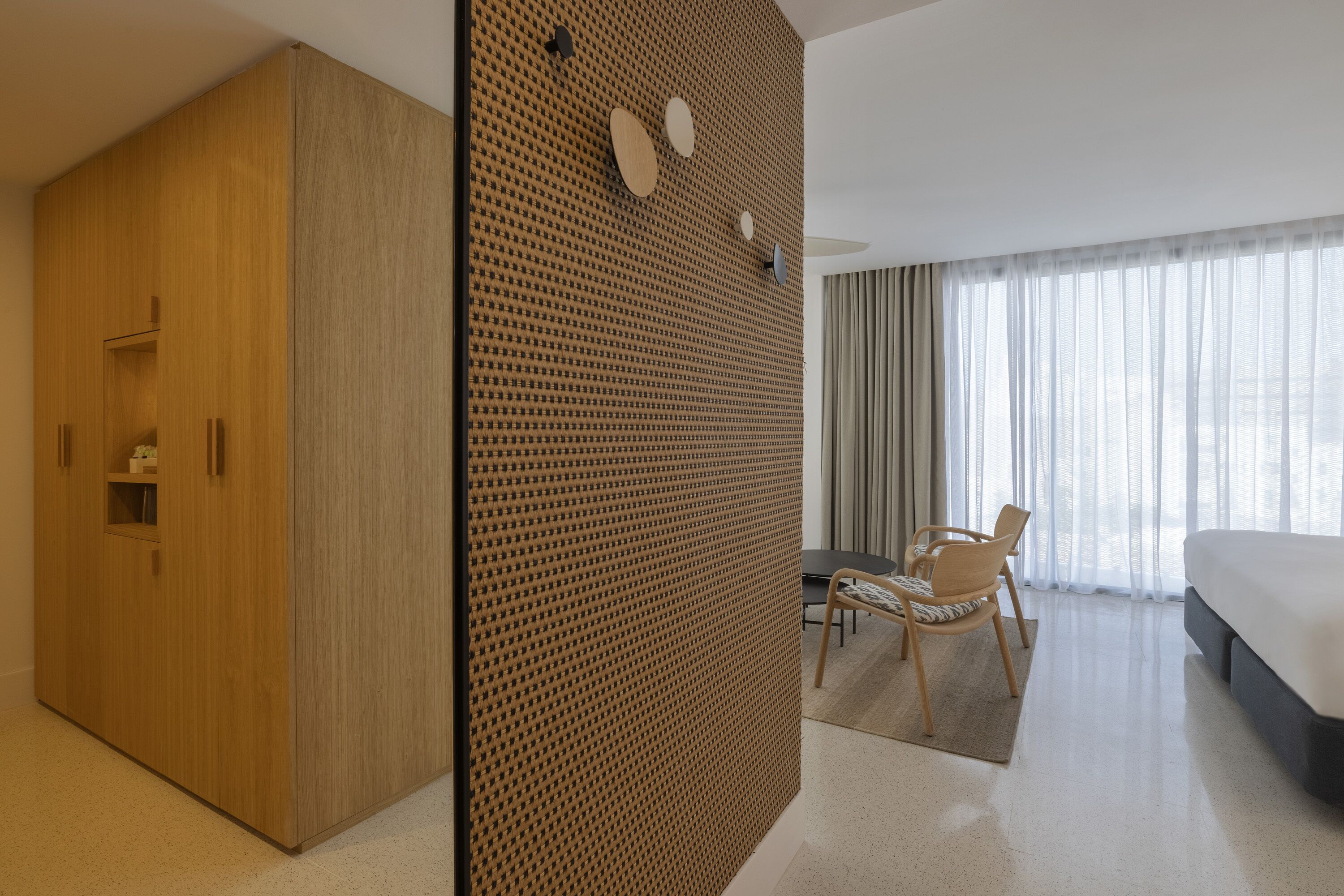 A hotel room with a large wardrobe, a wooden partition with circular cutouts, a dining area with a table and chairs, and a bed with a large window.