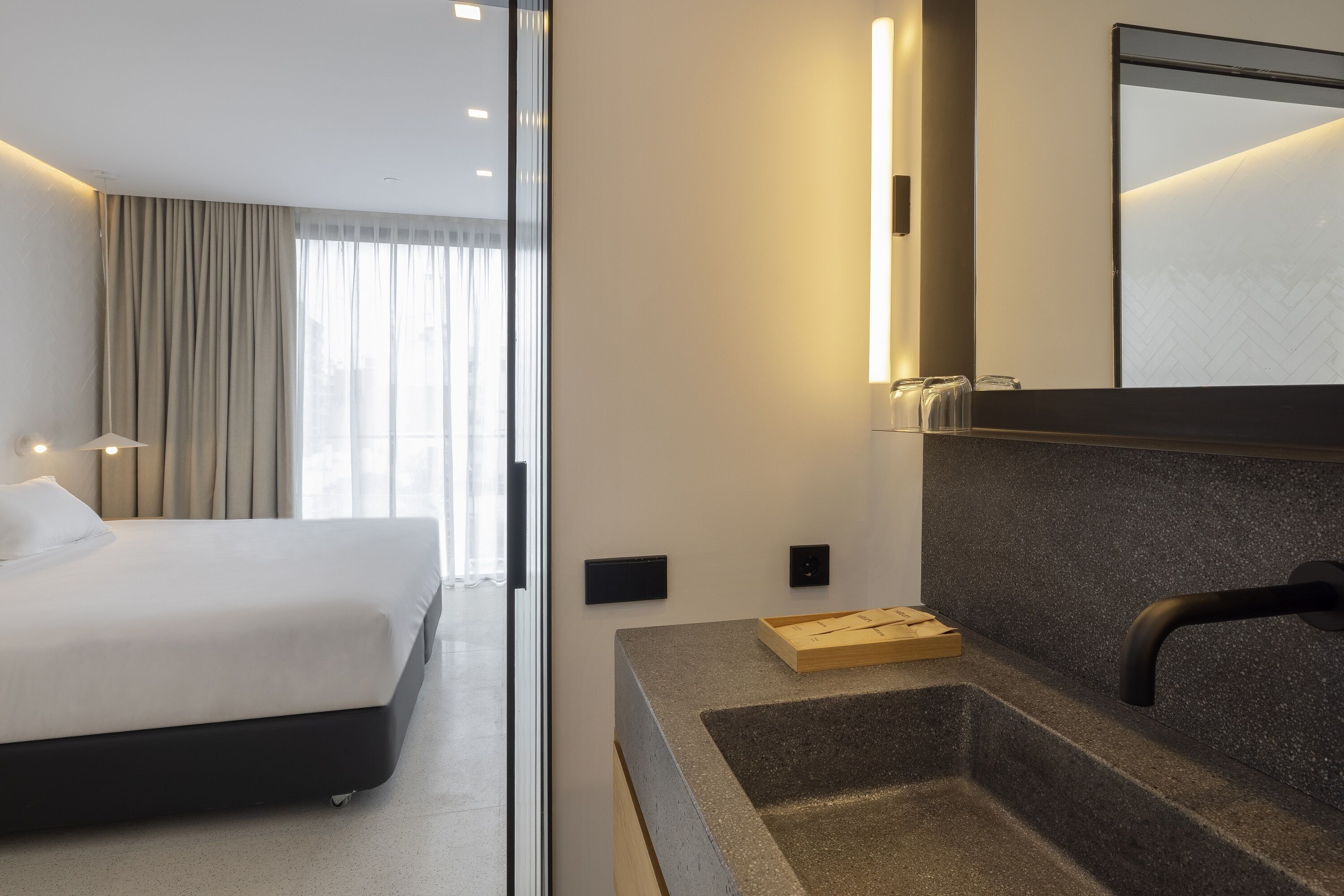 A modern hotel room with a bed, a vanity with a mirror, a sink, and a window with curtains.