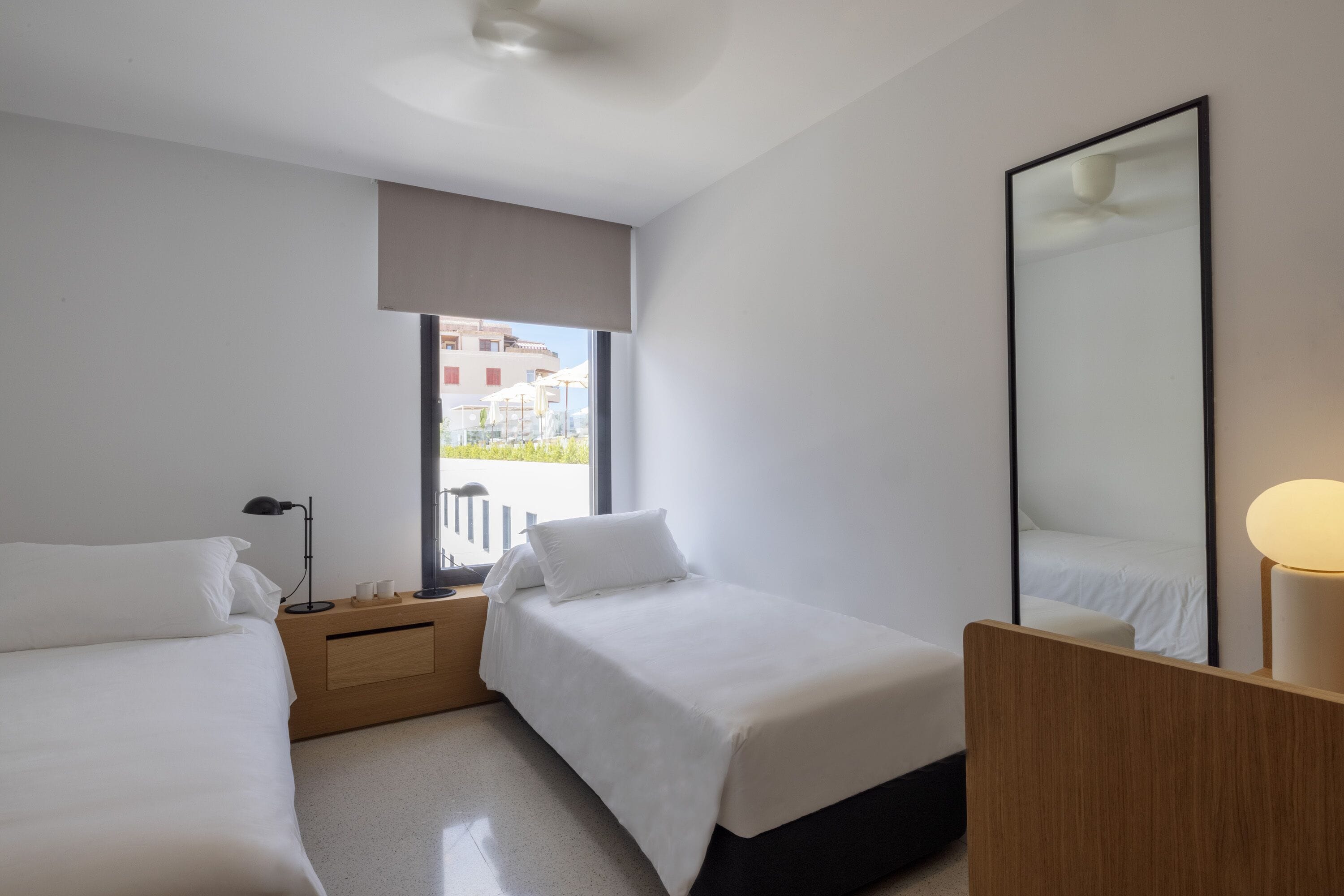 Habitación de hotel con dos camas, una mesita de noche de madera, un espejo y una ventana con vistas a edificios.