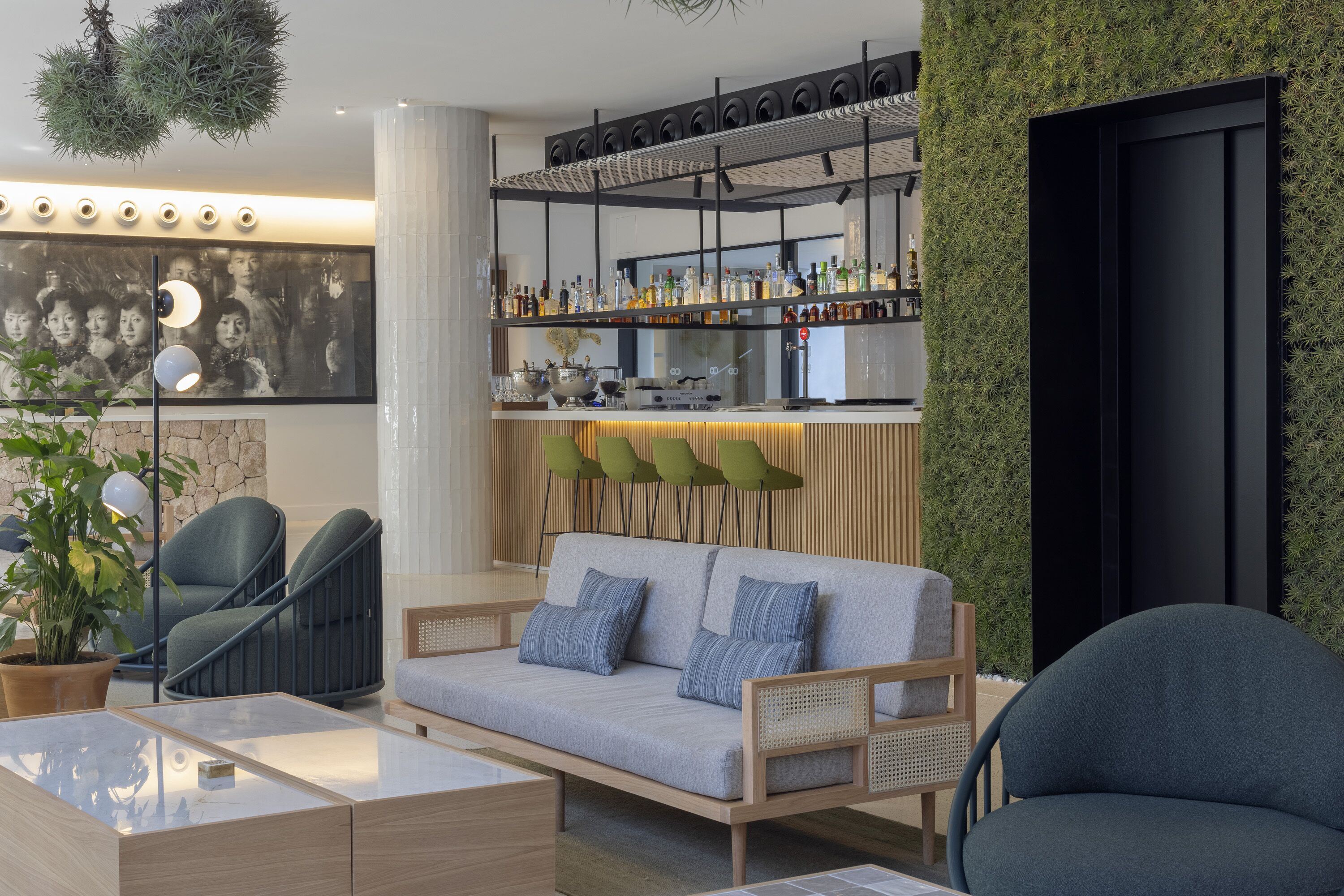 Uma área de estar moderna com sofá, cadeiras, um bar com prateleiras cheias de garrafas e uma parede com plantas.
