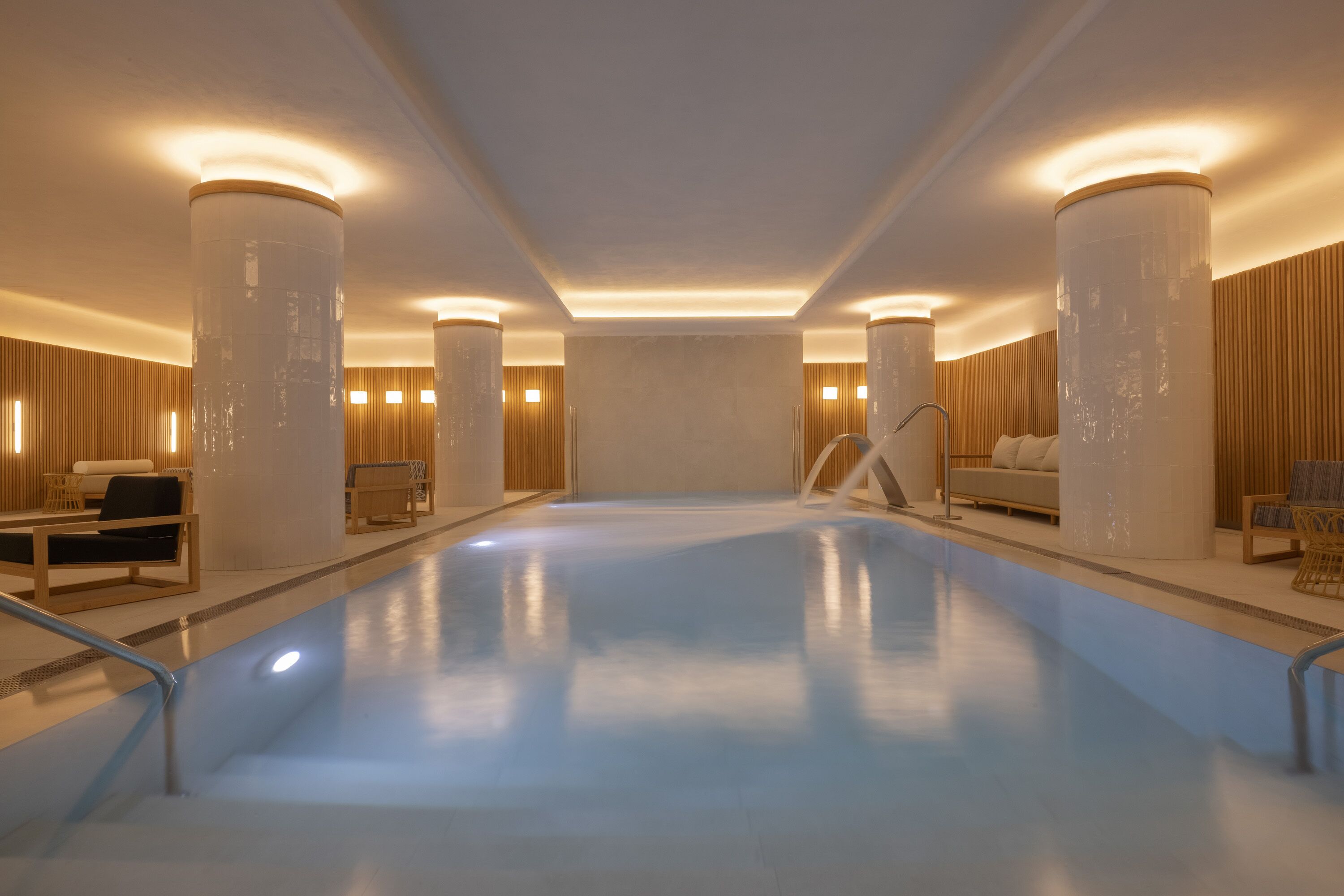 A spacious indoor swimming pool with a clear blue water surface, surrounded by wooden panel walls and modern seating areas.