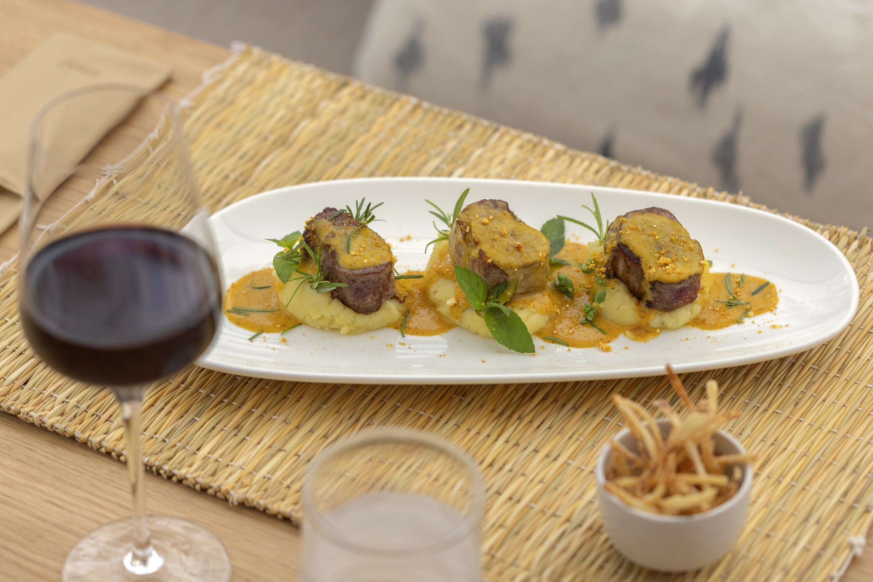 Une assiette blanche contenant de la nourriture et un verre de vin rouge, posés sur une table recouverte d’un set de table en fibres tressées.