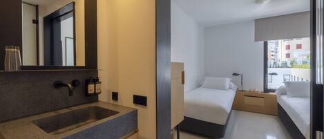 A modern hotel room with a large mirror, a granite sink, two beds, a bedside table with lamps, and a view of the city through the window.