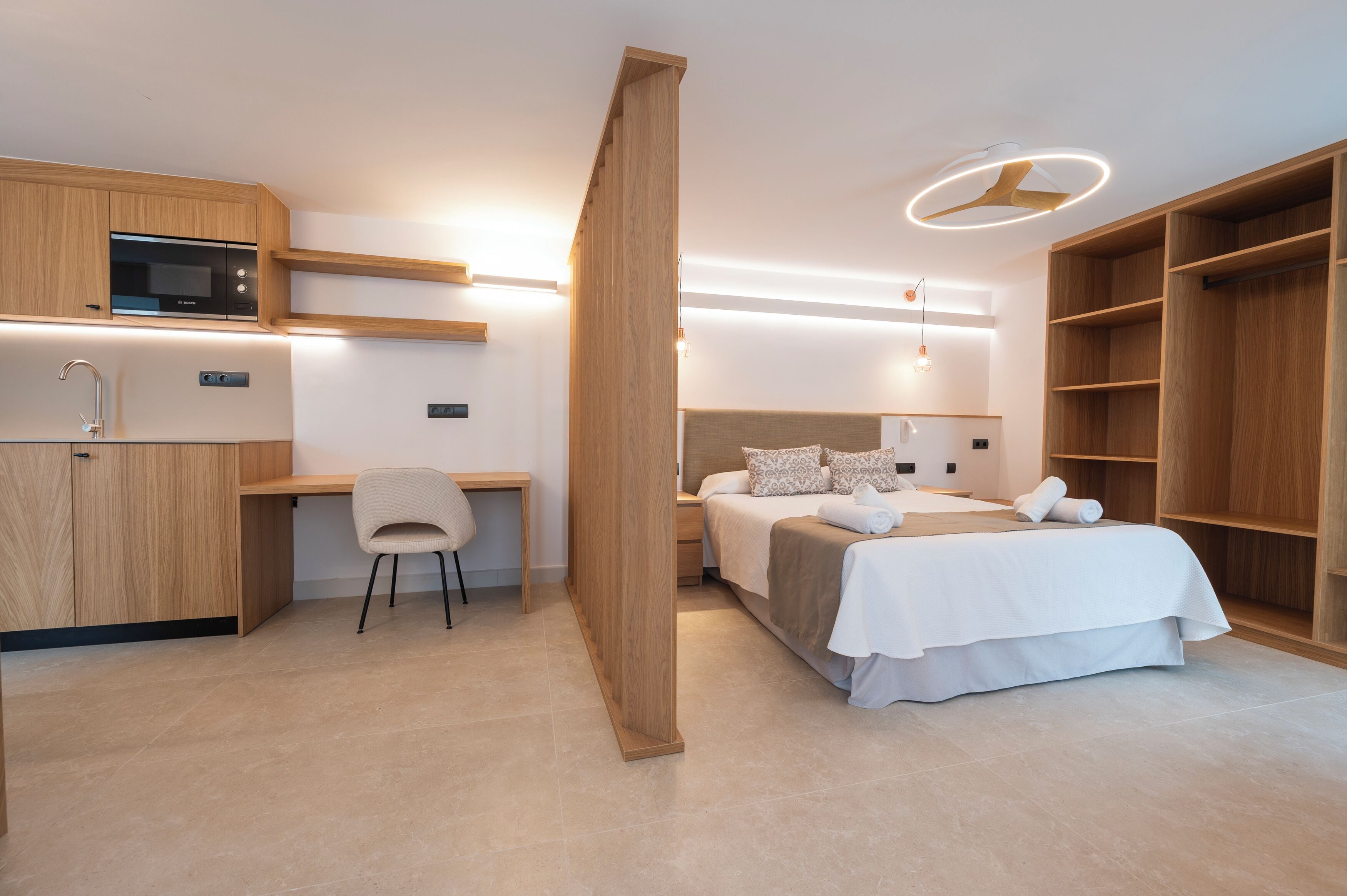 Una habitación de hotel moderna con kitchenette, una cama con ropa blanca, un escritorio con silla y estanterías empotradas de madera.