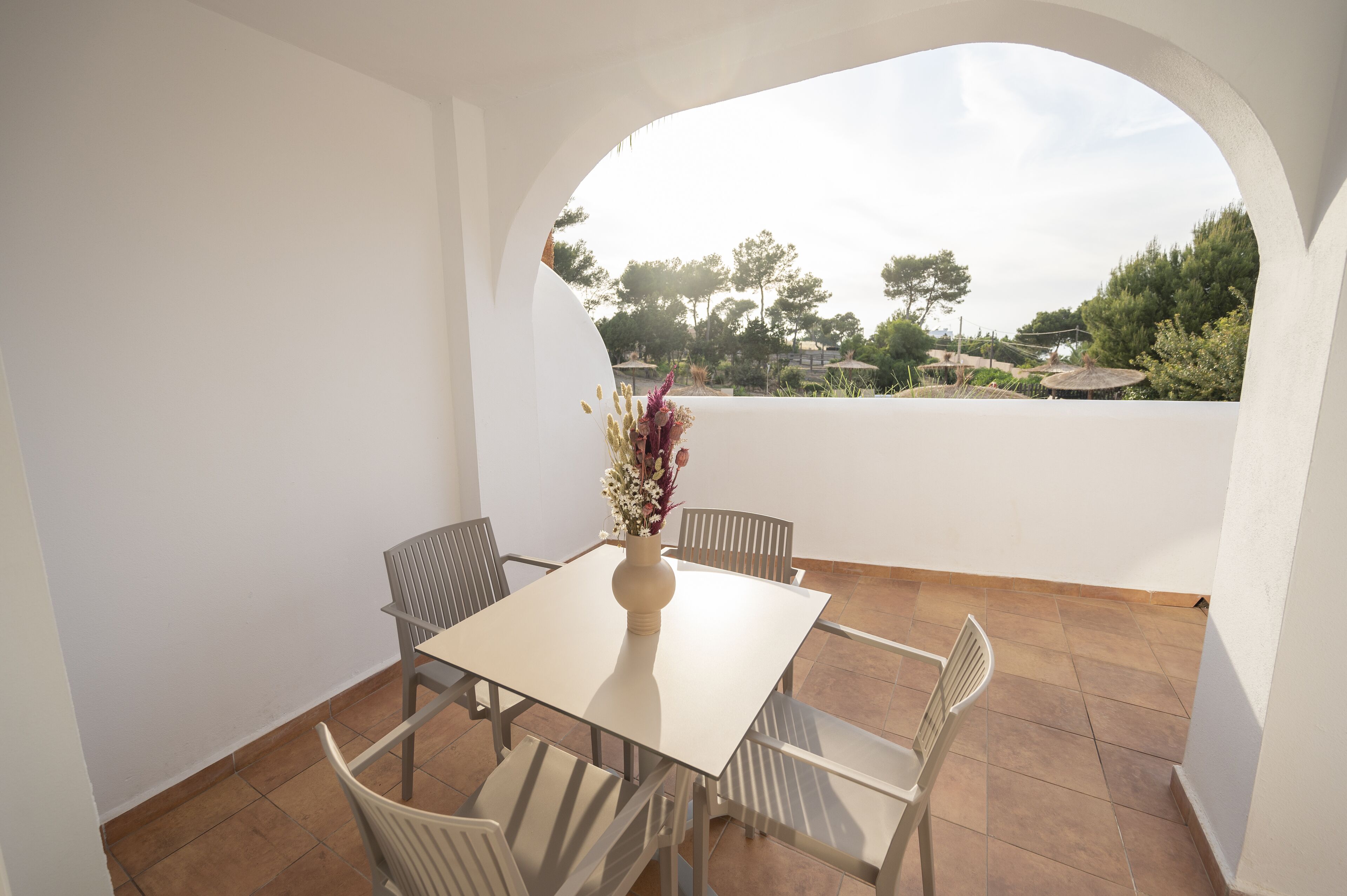 Comedor con mesa y sillas, un jarrón con flores y vistas a árboles y un edificio a través de un arco.