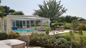 A covered outdoor swimming pool with a glass enclosure, surrounded by a garden with various plants and flowers. - Comfortable house with swimming pool in season for 8 people close to all amenities (Amboise)