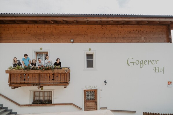 A building with a wooden balcony, a white wall with 'Gogger Hof' written on it, a small window, and a door with a decorative glass panel.