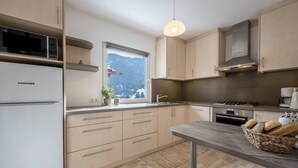 Moderni keittiö, jossa on puukalusteet, sisäänrakennettu mikroaaltouuni, ikkuna, josta avautuu näkymä vuorille, sekä ruokailutila, jossa on pöytä ja leipäkori.