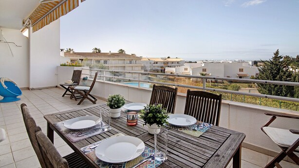 A balcony with a table set for dining, chairs, a small potted plant, and a view of buildings and a swimming pool. - 'Vilamoura Apartment 3 bedroom' with Sea View, Terraces, Shared Pool & Wi-Fi (Vilamoura)
