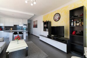 A modern living room with a flat-screen TV, a dining area with chairs, a kitchen with white cabinets, and a yellow accent wall.