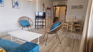 Un salon avec un canapé blanc, deux fauteuils en osier avec des coussins bleus, un téléviseur posé sur un meuble en bois, un coin repas avec des chaises et une table en bois, et des œuvres d’art accrochées aux murs.