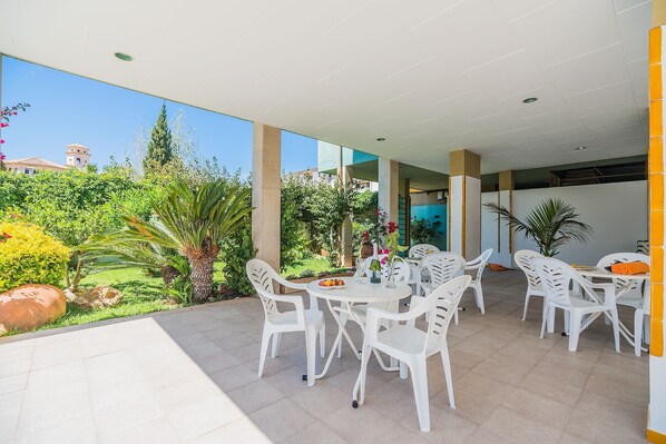 An outdoor patio with white plastic chairs and tables, surrounded by greenery and a view of buildings in the distance.