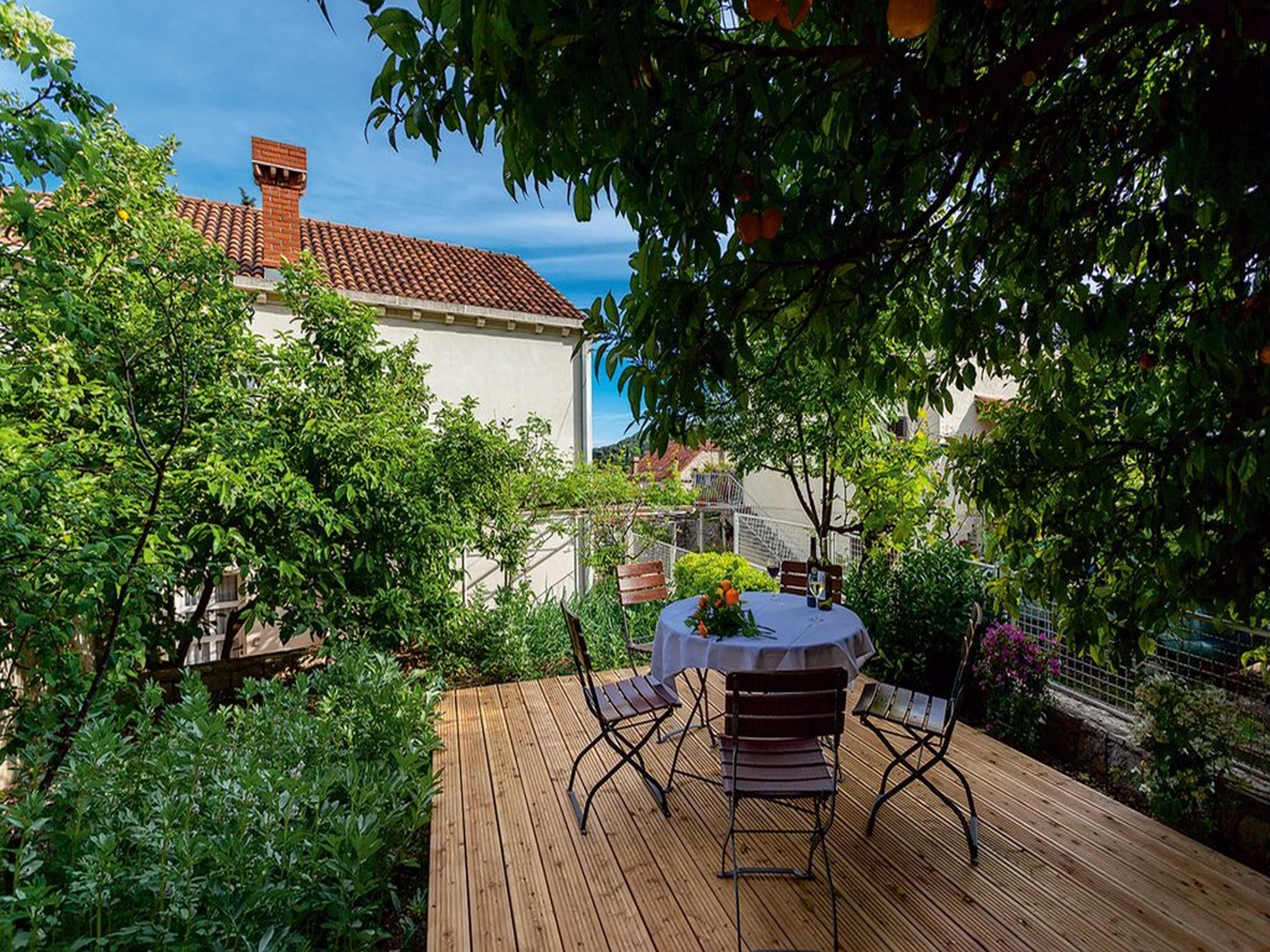 A wooden deck with a round table and two chairs, surrounded by lush greenery and a building with a red-tiled roof.