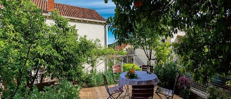 A wooden deck with a round table and two chairs, surrounded by lush greenery and a building with a red-tiled roof.