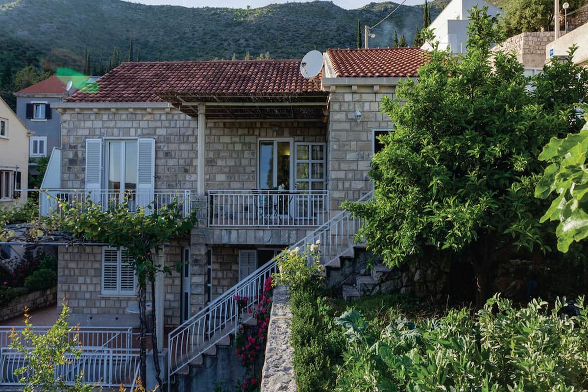 A stone house with a red-tiled roof, a balcony with white railings, and a view of a mountainous landscape.