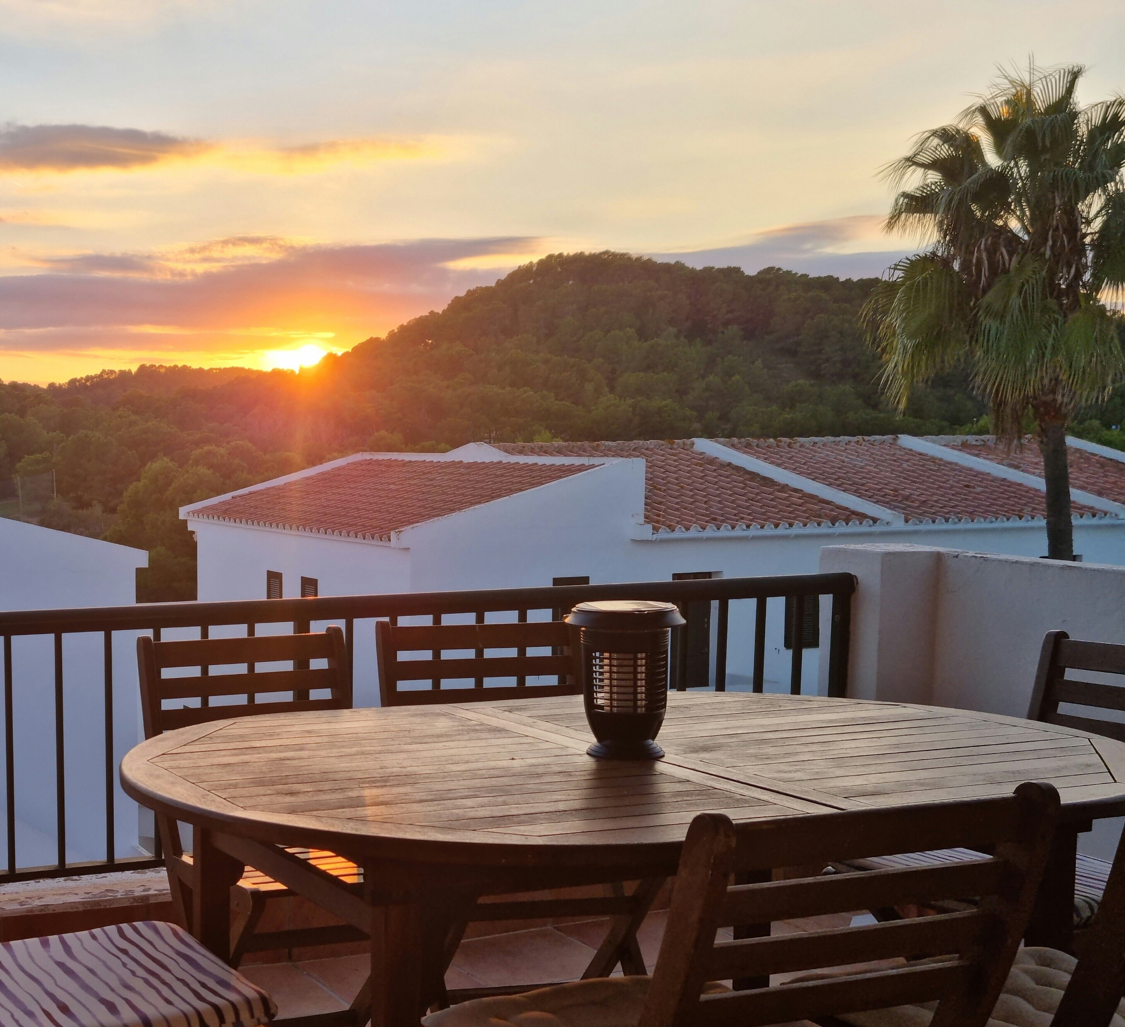 Mesa redonda de madera con sillas y una lámpara de mesa en un balcón con vistas a una puesta de sol y un paisaje montañoso.