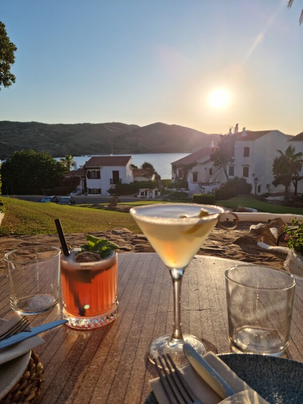 A table with drinks and cutlery, overlooking a body of water and buildings.
