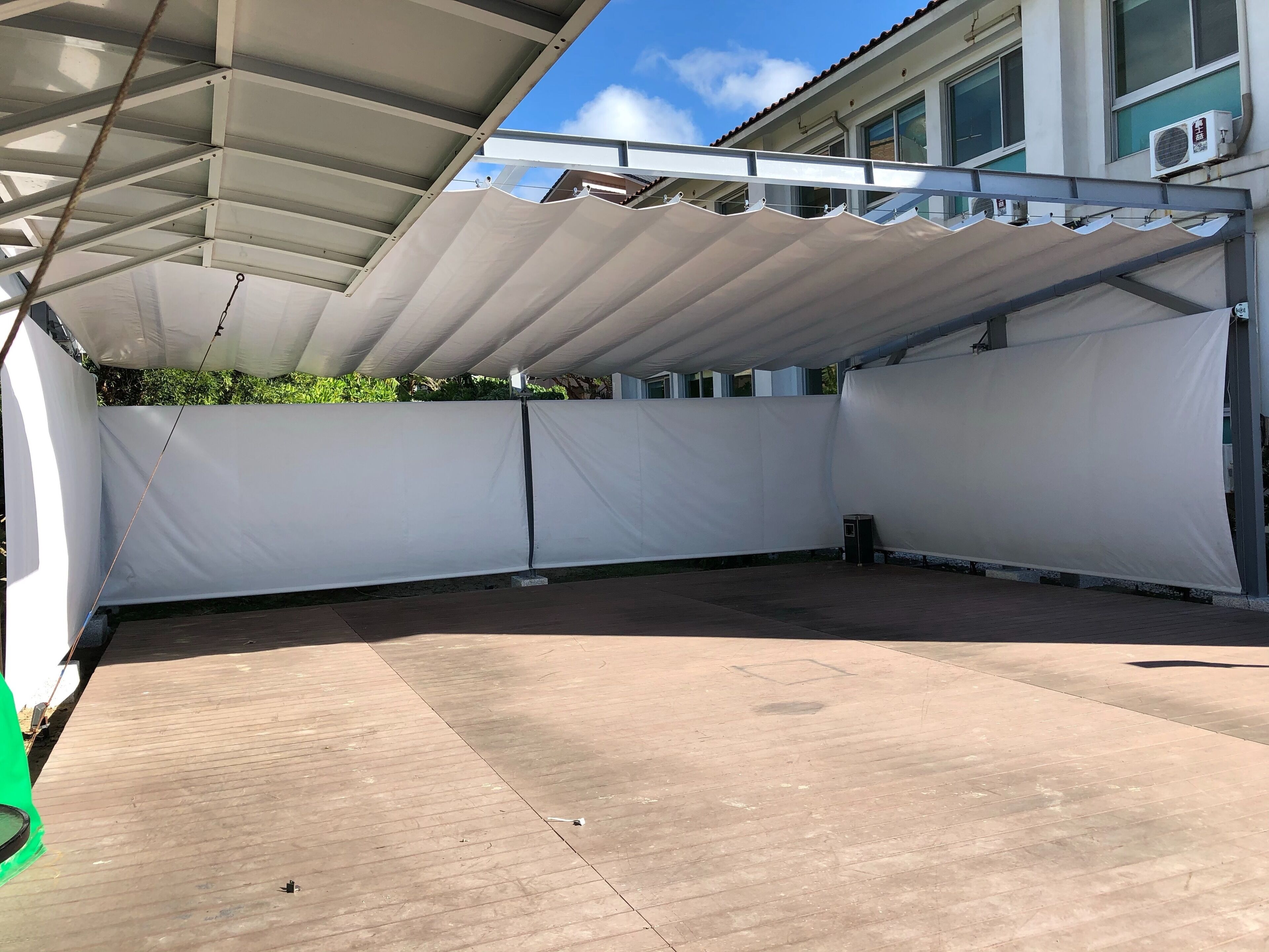 Un espace extérieur abrité doté d’un sol en béton, de stores blancs rétractables et d’un bâtiment avec de grandes fenêtres en arrière-plan.