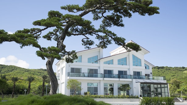 Un edificio moderno de dos pisos con amplios ventanales y un balcón de vidrio, rodeado de exuberante vegetación y un árbol destacado.