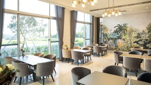 Un restaurante con varias mesas y sillas, ventanas amplias con cortinas, y un mural de plantas tropicales en la pared.