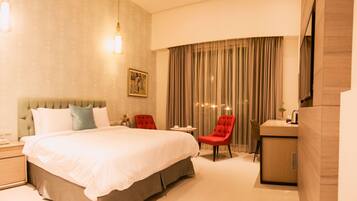 A hotel room with a large bed, a red chair, a small table, and a desk with a mirror.